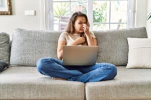 Beautiful young brunette woman sitting on the sofa using computer laptop at home smelling something stinky and disgusting, intolerable smell, holding breath with fingers on nose. Septic System Odor - Red Dirt Septic